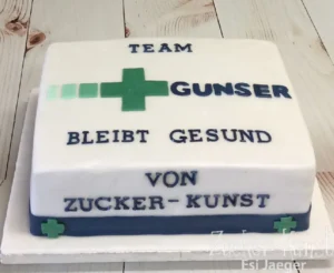 Rechteckige weiße Torte mit Aufschrift "Team Gunser Bleibt Gesund", verziert mit einem grünen Kreuz und einem dunkelblauen Band.