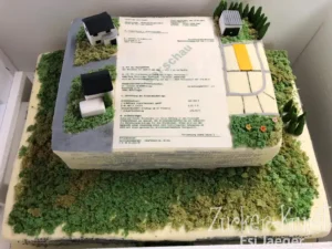 Zweistöckige Torte in Form eines Grundstücks mit Gebäuden, Straßen und einem Foto-Druck eines Vertrags oder Bauplans.