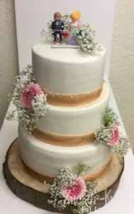 Rustikale dreistöckige Hochzeitstorte: Weißer Fondant, Holzscheiben-Unterlage, echte Blumen und Playmobil-Brautpaar. Zucker-Kunst Esi Jaeger.