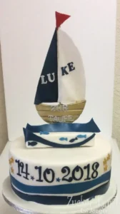 Zweistöckige Tauftorte in Blau und Weiß, gekrönt von einem Segelboot, mit Fisch-Motiven und dem Datum der Taufe.