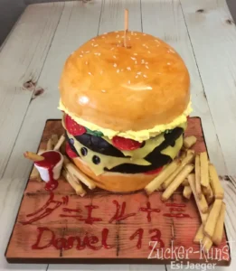 Realistisch modellierte Burger-Torte, angerichtet auf einem Holzbrett mit Ketchup und Pommes-Attrappen.