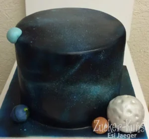Hohe, runde Torte in tiefem Dunkelblau mit türkisfarbenen Sprenkeln, die eine Galaxie darstellen, verziert mit Modellen von Erde, Mond und einem Planeten.