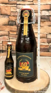 Sehr große, dunkelbraune Torte in Form einer Bügelverschluss-Bierflasche, dekoriert mit einem 60. Geburtstag-Etikett neben dem Original.